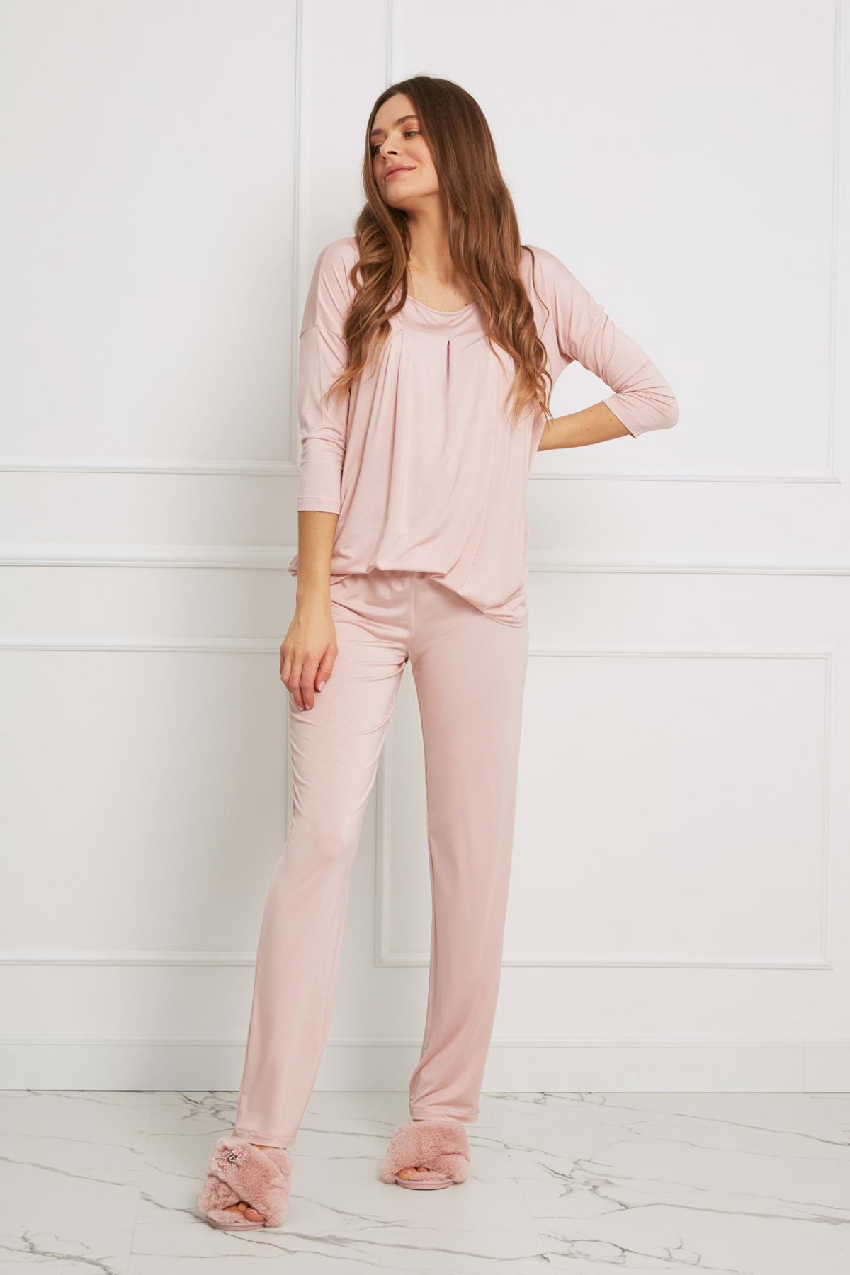Kobieta z długimi brązowymi włosami stoi pod białą ścianą, ubrana w Loungewear luźny komplet z długimi spodniami - brudny róż i pasujące do niego rozmyte kapcie. Opiera jedną rękę na biodrze i spogląda w bok ze zrelaksowanym wyrazem twarzy.