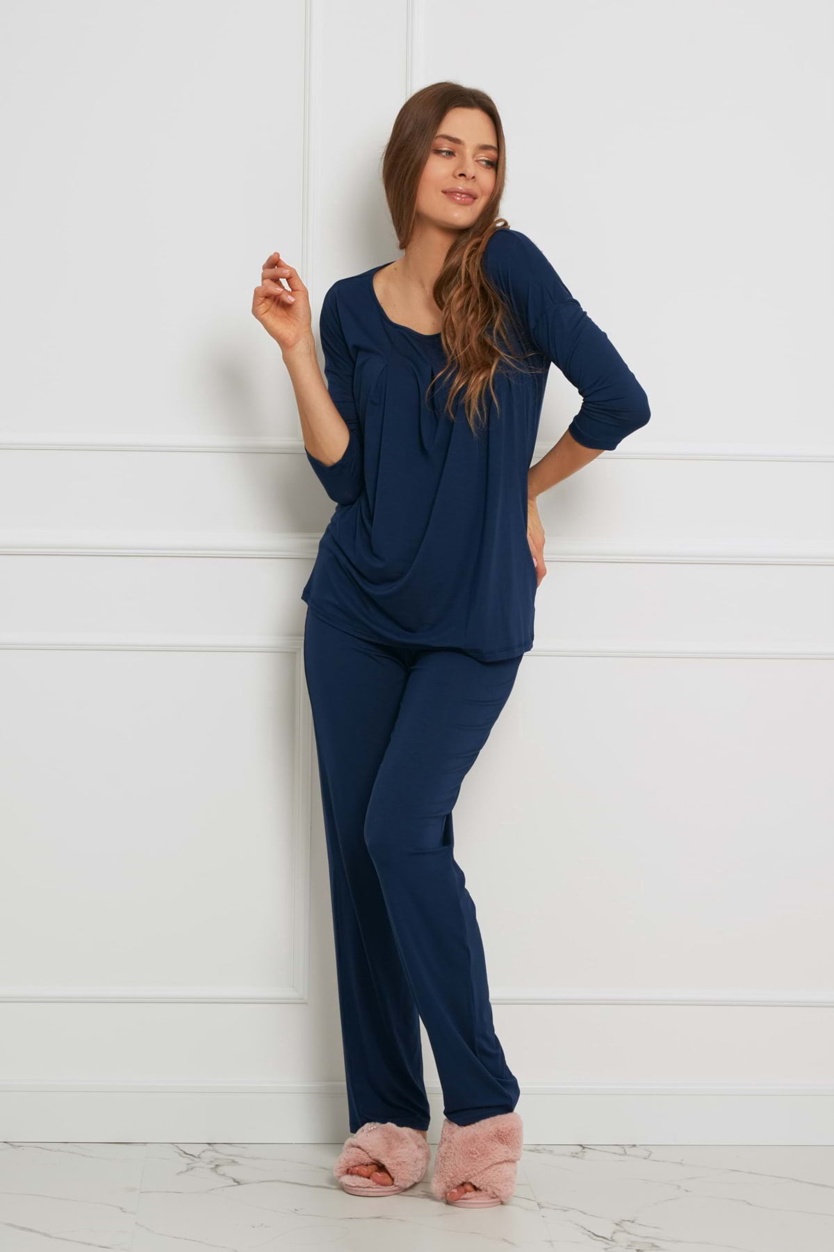 Kobieta stoi w pomieszczeniu pod białą ścianą, ubrana w Loungewear luźny komplet z długimi spodniami - granatowy i różowe puszyste kapcie. Ma długie brązowe włosy, uśmiecha się i unosi jedną rękę blisko twarzy.