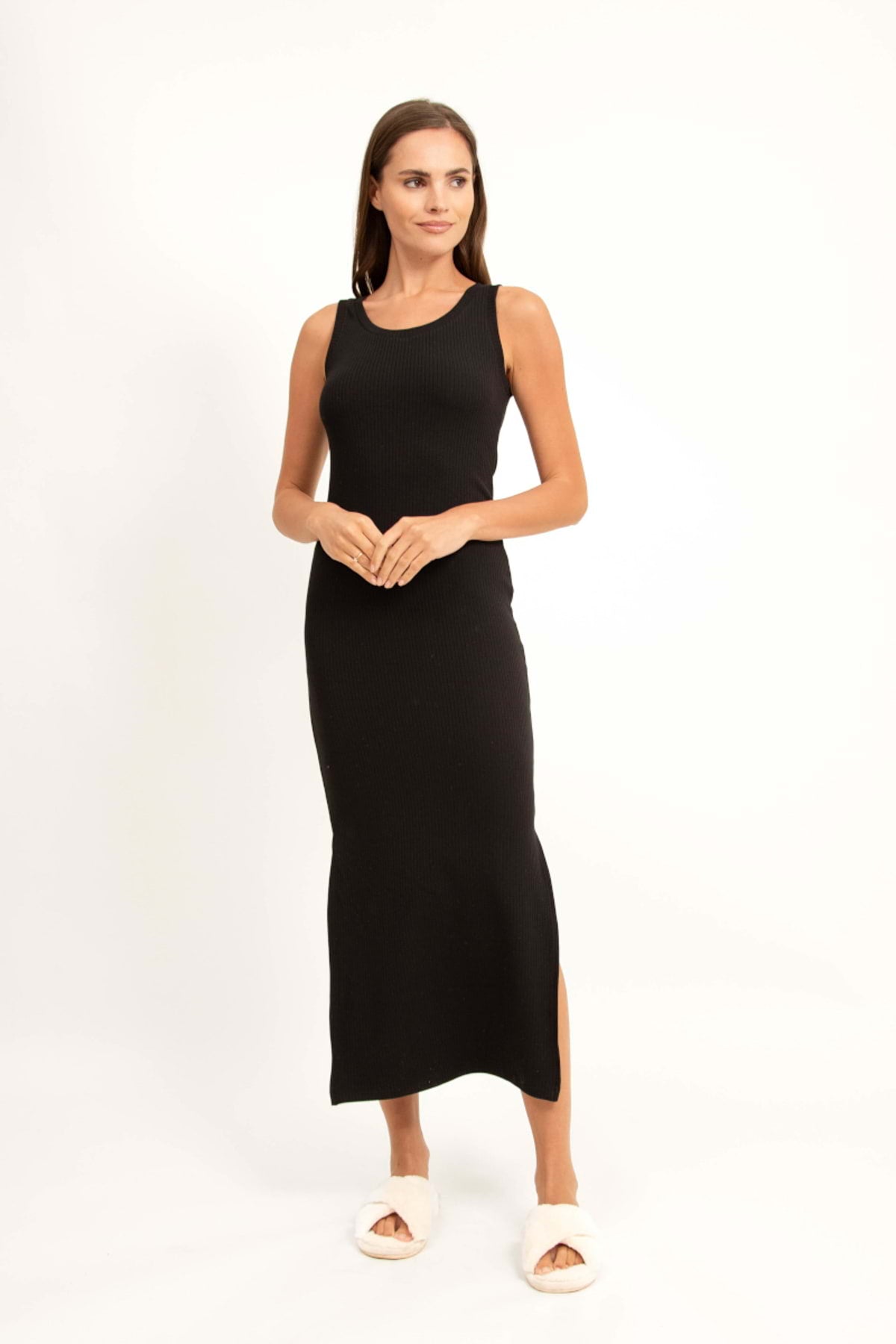 Kobieta o prostych brązowych włosach stoi na białym tle, ubrana w czarną loungewear wiskozową sukienkę maxi bez rękawów i białe pantofle. Spogląda lekko w bok, delikatnie uśmiechając się z dłońmi splecionymi z przodu.