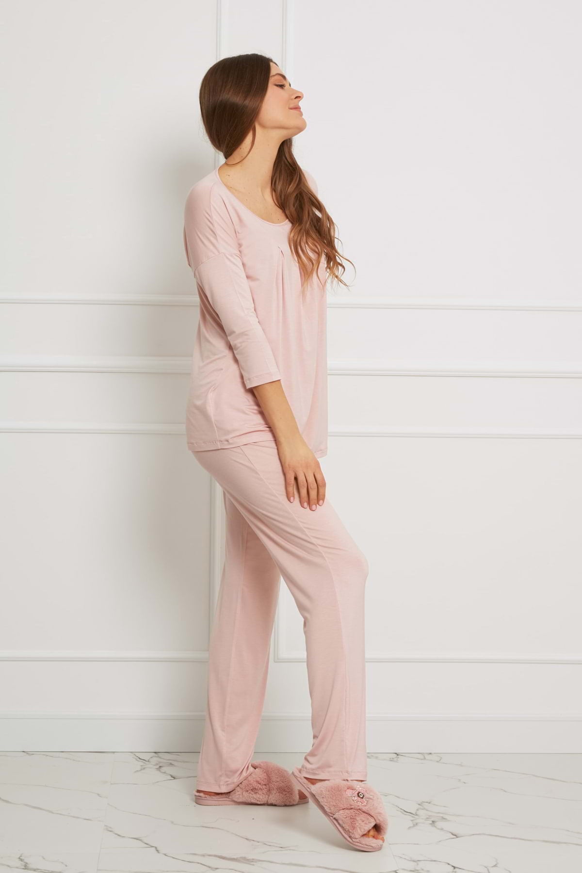 Kobieta z długimi, falującymi włosami pozuje z profilu na tle białej ściany, ubrana w Loungewear luźny komplet z długimi spodniami - brudny róż i pasujące rozmyte kapcie na marmurowej podłodze.