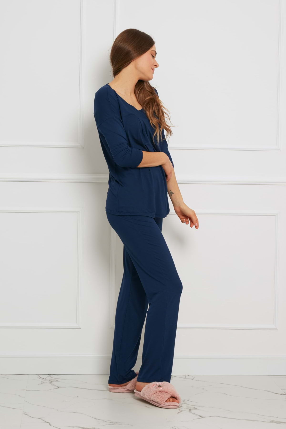 Kobieta z długimi brązowymi włosami ma na sobie Loungewear luźny komplet z długimi spodniami - granatowy i różowe kapcie, stojąc bokiem do białej ściany wyłożonej panelami i marmurowej podłogi, patrząc w dół z rękami rozluźnionymi u boku.