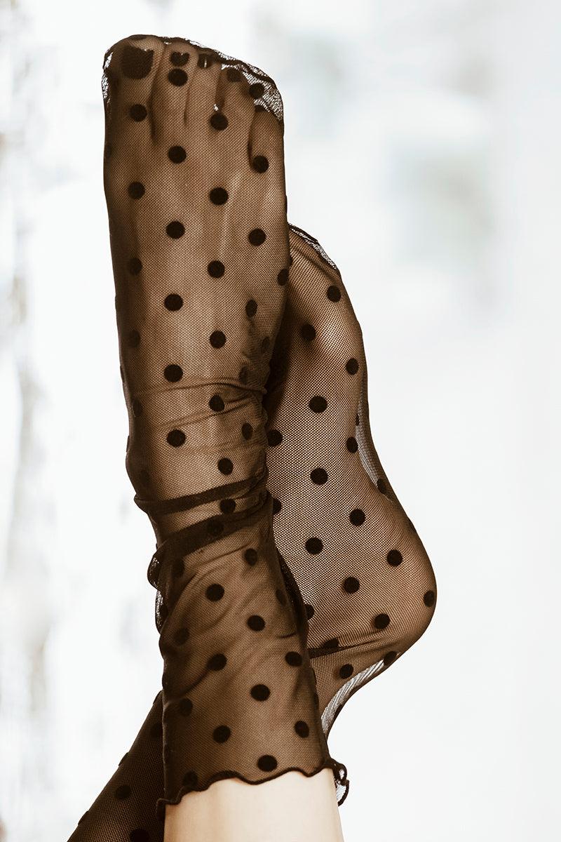 Zbliżenie na dwie stopy ubrane w Tiulowe skarpetki Czarne Retro z wzorem w kropki. Miękko rozmyte tło podkreśla ich delikatną fakturę - idealne do eleganckich stylizacji.