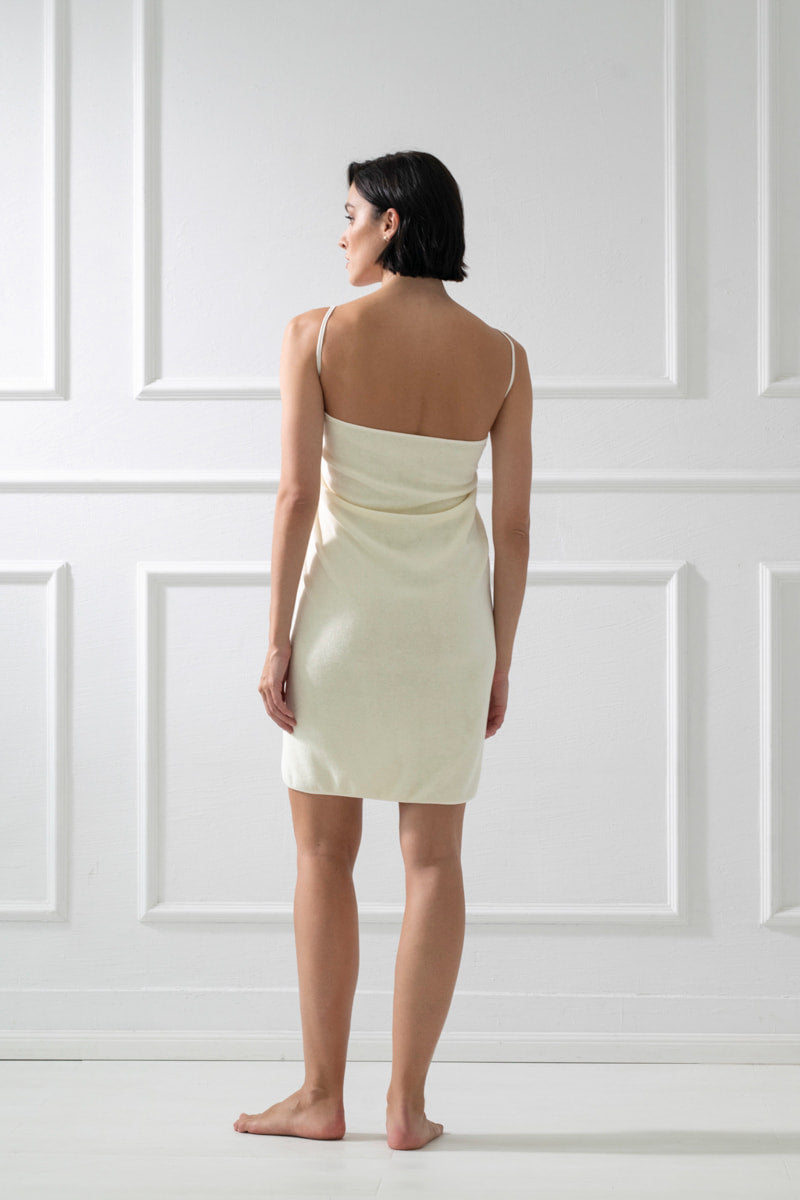 Kobieta o krótkich ciemnych włosach, bosa i odwrócona tyłem, ma na sobie jasnokremową halkę i Calma uniwersalna chusta bawełniana na ramiączkach - waniliowa w jasnym, minimalistycznym pokoju z białymi panelami na ścianach.